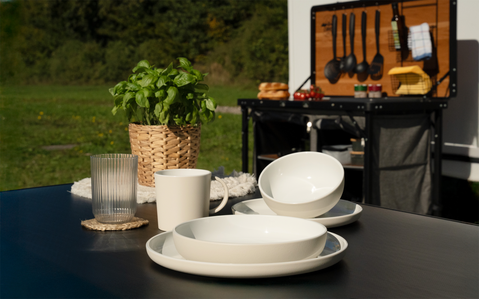 Table noire avec assiettes et bols blancs empilés, un verre, une tasse, une plante verte en pot dans un panier tressé ; en arrière-plan, une boîte de cuisine ouverte avec des ustensiles de cuisson suspendus et une planche en bois