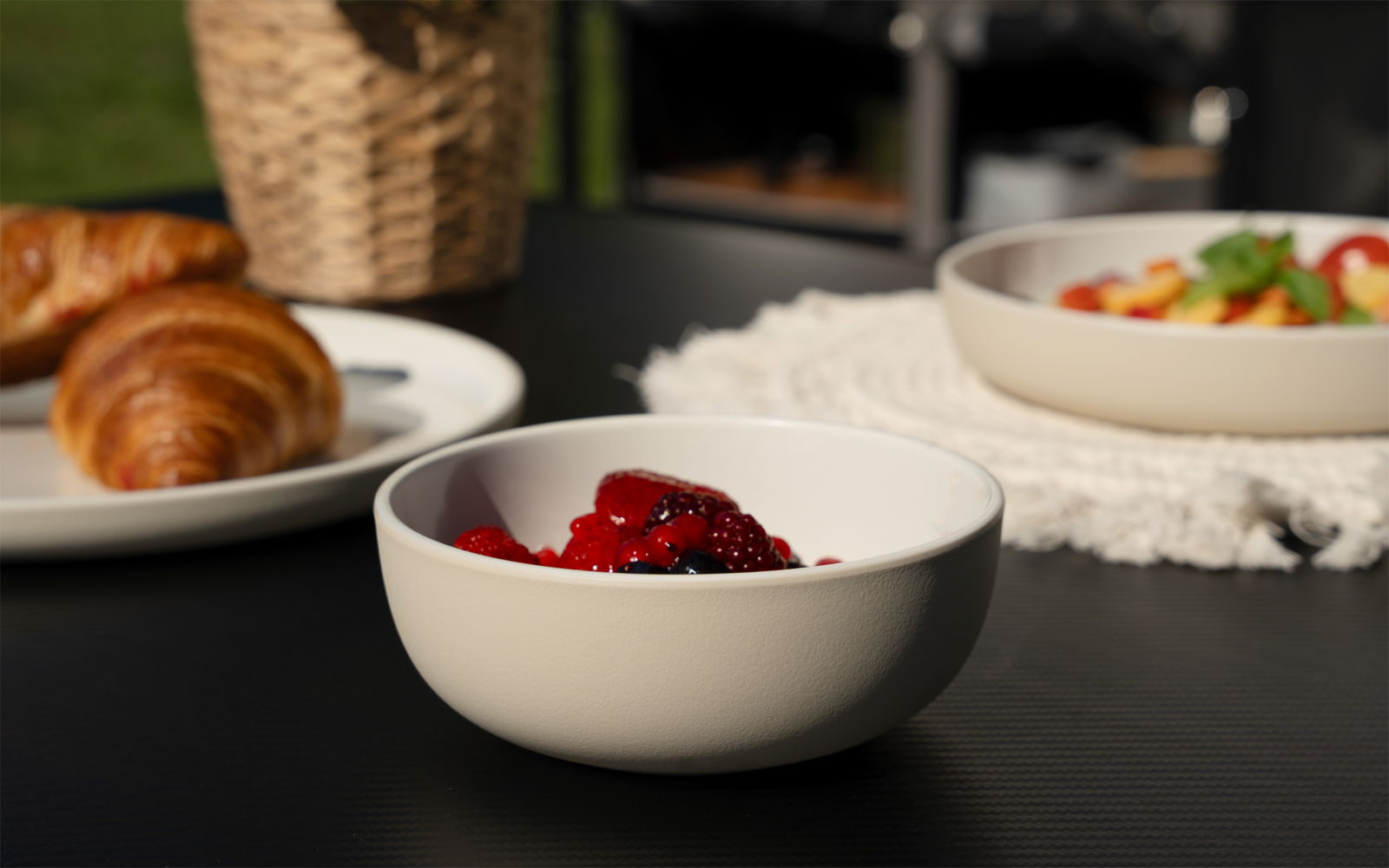 Bol blanc avec des fruits rouges sur une table foncée, en arrière-plan des croissants sur une assiette, un panier en osier et un bol de fruits coupés
