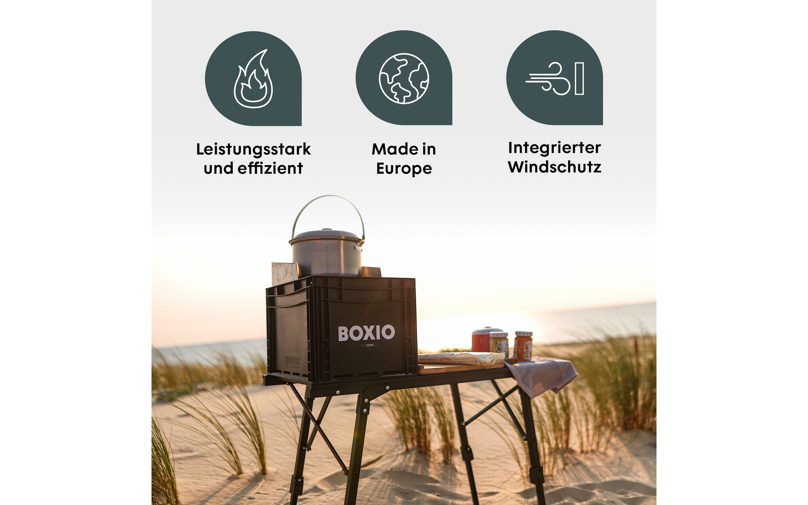 Réchaud portable noir marqué BOXIO sur une table pliante sur une plage de sable ; casserole argentée sur le réchaud, deux boîtes sur la table ; en haut trois icônes rondes vertes avec symboles blancs et texte allemand