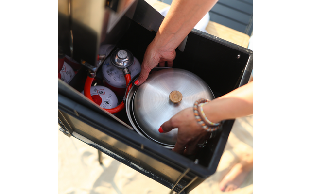Mains avec vernis à ongles rouge et bracelet de perles tiennent un couvercle rond en acier inoxydable avec bouton en bois dans une boîte noire ouverte; la boîte contient plusieurs cartouches de gaz avec raccords rouges et un tuyau rouge, sol sablonneux en arrière‑plan