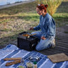 Personne aux cheveux roux en chignon, portant un sweat-shirt bleu et un jean, agenouillée sur une planche en bois à côté d'une caisse noire avec un petit réchaud, remuant une casserole. Devant elle une nappe de pique-nique bleu et blanc avec deux baguettes, un verre, un bol en métal, une planche à découper et des pots; en arrière-plan herbe et colline