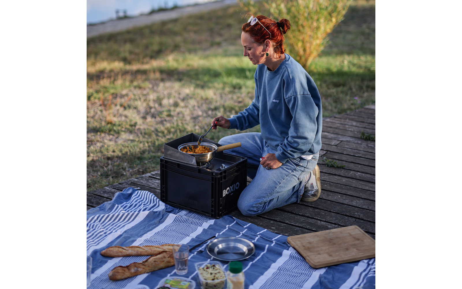 Personne aux cheveux roux en chignon, portant un sweat-shirt bleu et un jean, agenouillée sur une planche en bois à côté d'une caisse noire avec un petit réchaud, remuant une casserole. Devant elle une nappe de pique-nique bleu et blanc avec deux baguettes, un verre, un bol en métal, une planche à découper et des pots; en arrière-plan herbe et colline