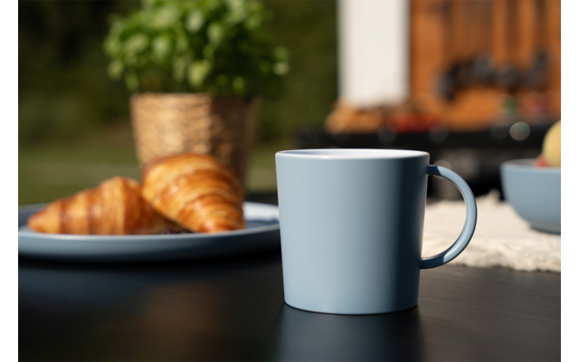 Hellblauer Keramikbecher im Vordergrund auf einem dunklen Tisch; unscharfe Platte mit zwei Croissants, Topf mit Basilikum und Schale mit Obst im Hintergrund