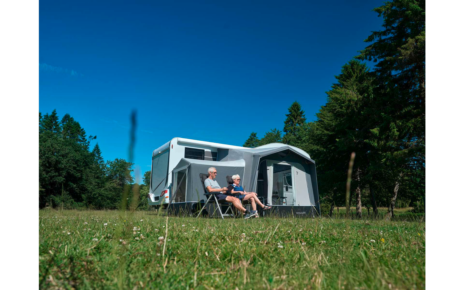 Zwei Personen sitzen auf Campingstühlen vor einem weißen Wohnwagen mit angebautem Vorzelt auf einer Wiese, im Hintergrund Bäume und blauer Himmel