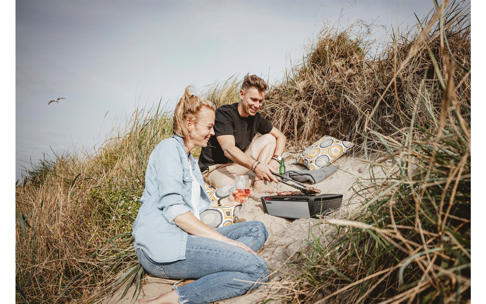 Zwei Personen sitzen auf einer Decke zwischen Sanddünen und Strandgras; auf der Decke liegen Picknickutensilien, eine Tasche und Getränke; im Hintergrund Himmel mit zwei fliegenden Vögeln