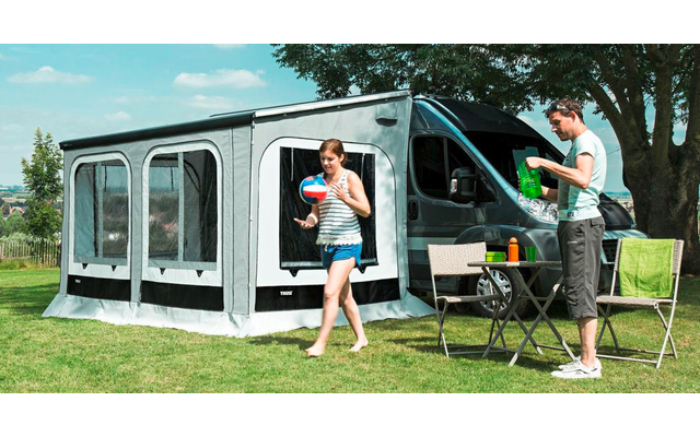 Wohnmobil mit angebautem Vorzelt auf einer Wiese, vor dem Zelt steht ein Tisch mit Campingstühlen, eine Frau und ein Mann sind draußen unterwegs