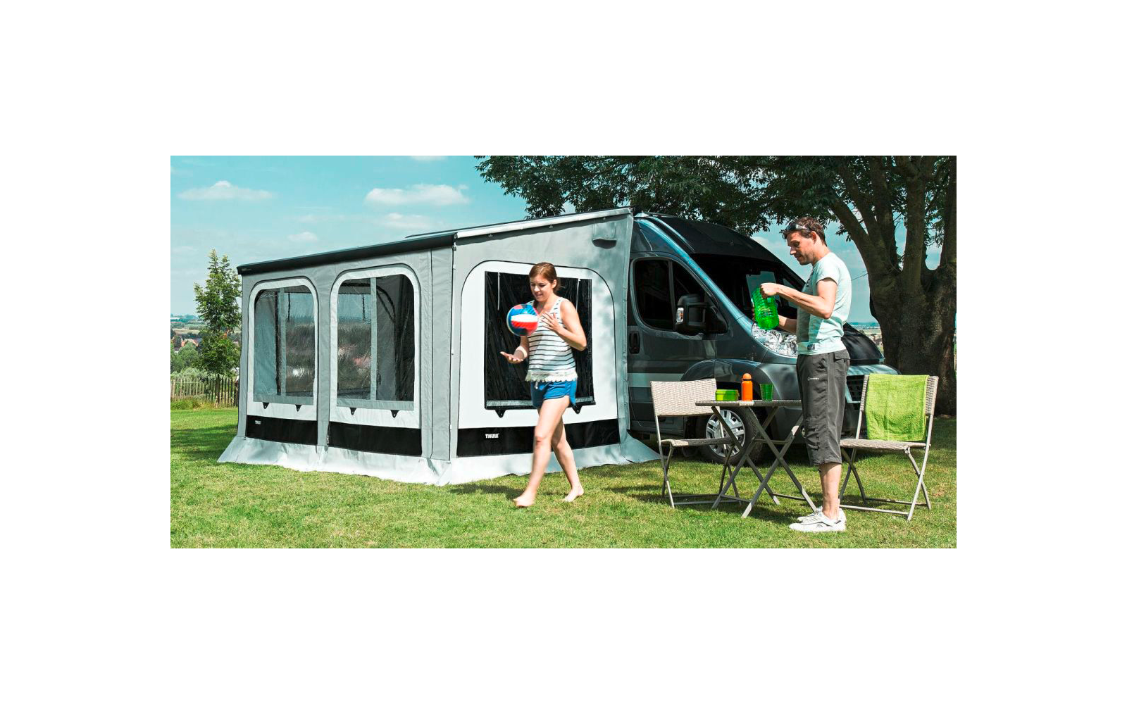 Wohnmobil mit angebautem Vorzelt auf einer Wiese, vor dem Zelt steht ein Tisch mit Campingstühlen, eine Frau und ein Mann sind draußen unterwegs