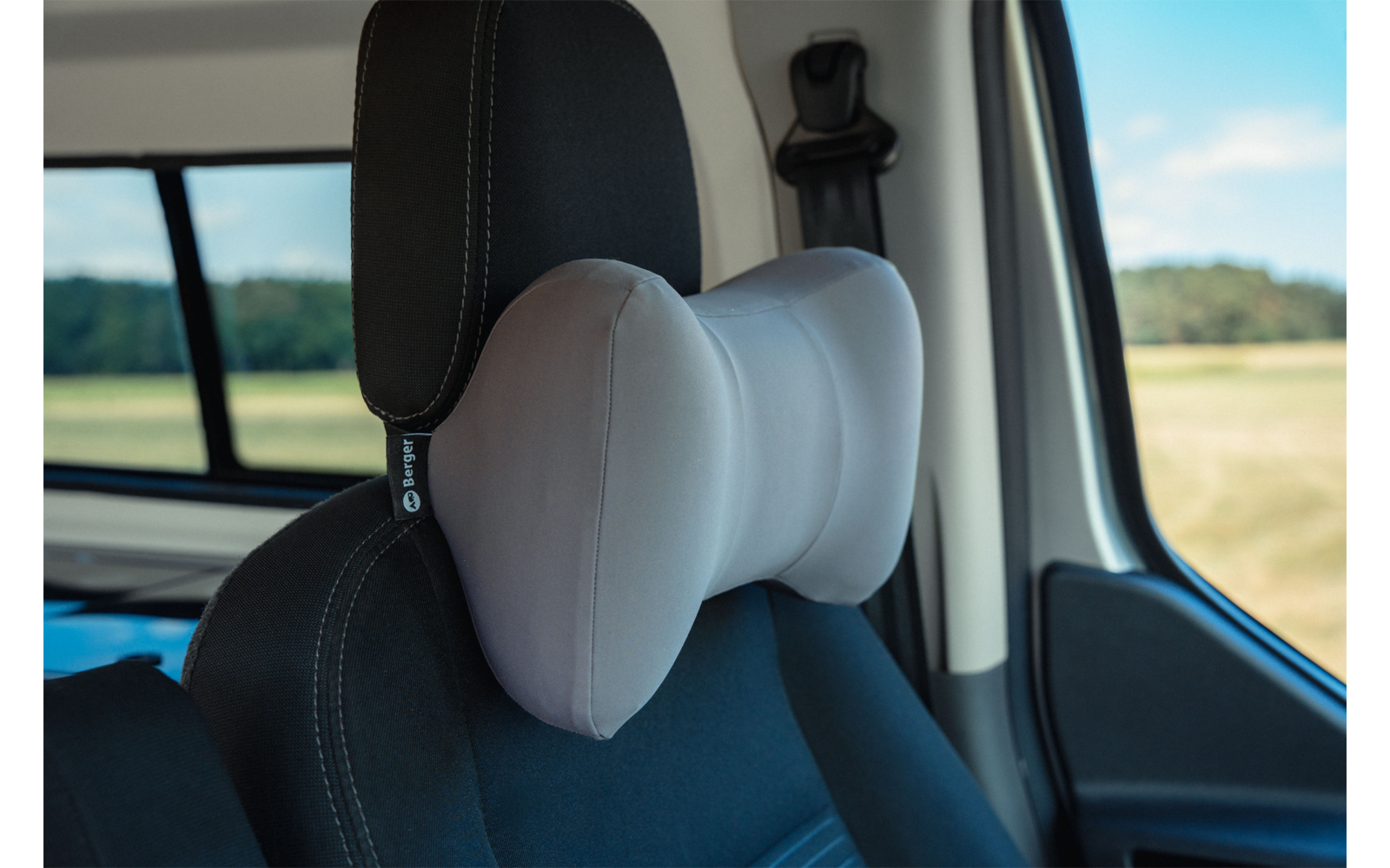 Gray butterfly-shaped neck pillow attached to the headrest of a black car seat; car window showing fields and blue sky
