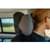 Person with blond hair tied back, wearing a black top and necklace, seated in a car; gray neck pillow attached to the headrest; window shows blurred green landscape