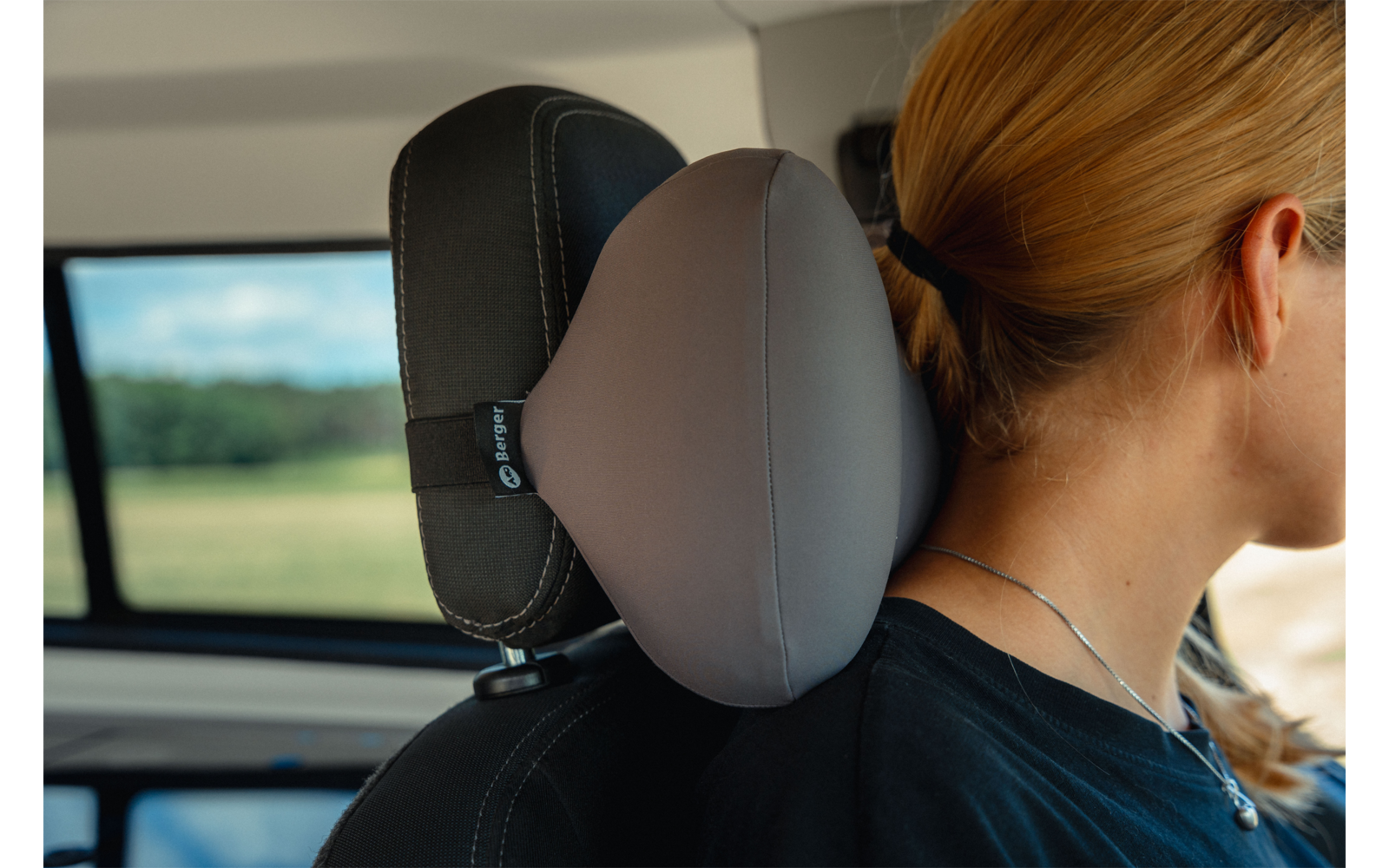 Person with blond hair tied back, wearing a black top and necklace, seated in a car; gray neck pillow attached to the headrest; window shows blurred green landscape