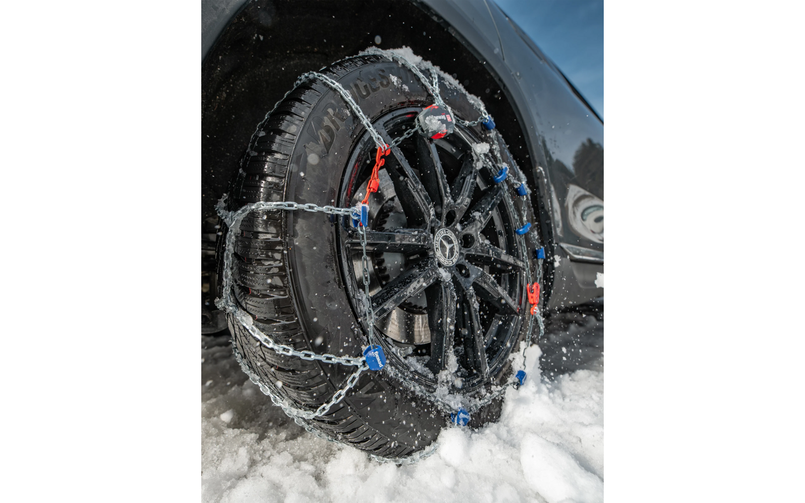 Plano detalle de un neumático de coche con cadenas para nieve en una llanta negra, nieve en el suelo y sobre las cadenas