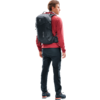 Person in roter Jacke und dunkler Hose mit schwarzem Rucksack, steht seitlich, Blick nach rechts