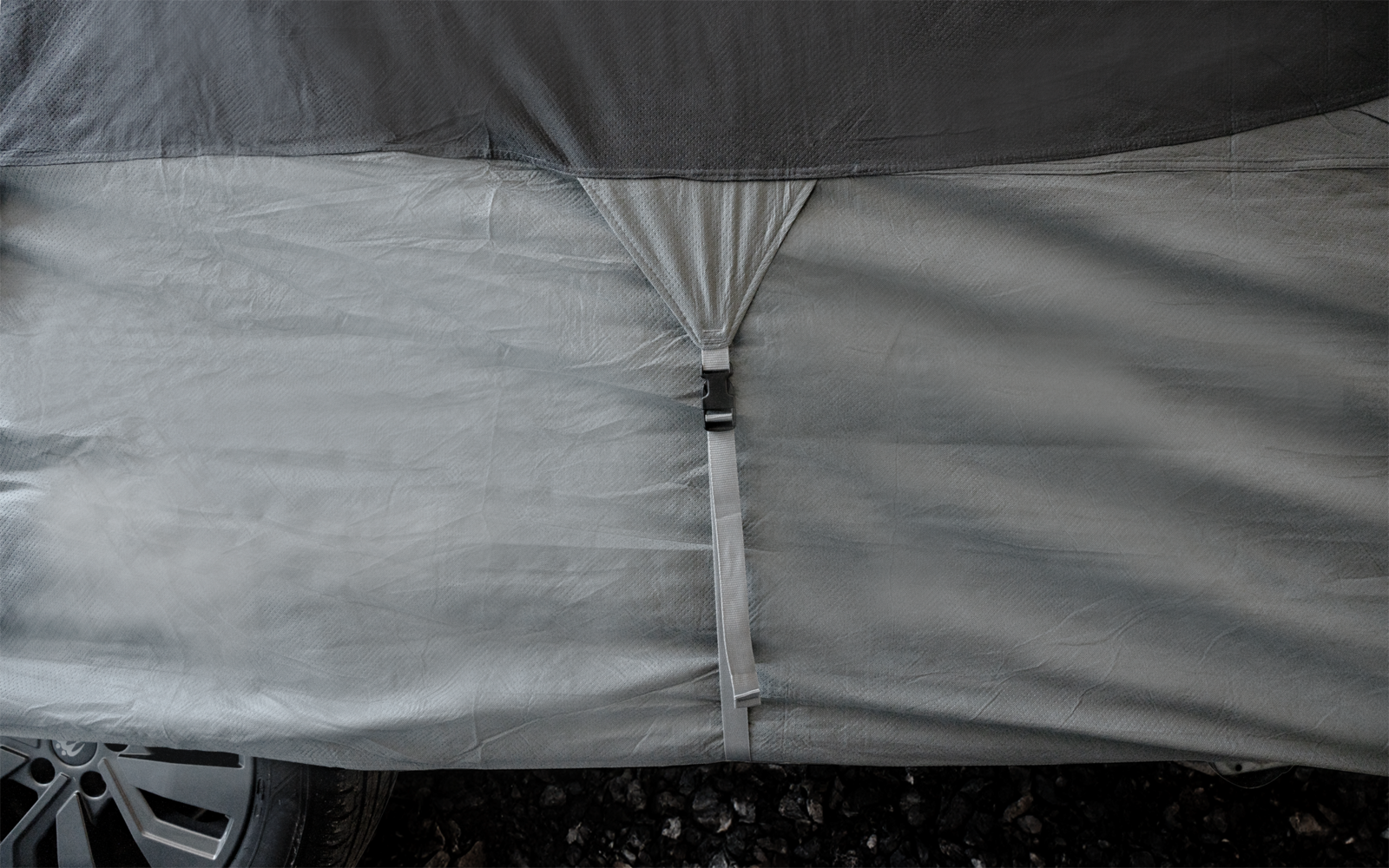 Gray cover with a central strap and buckle; part of a wheel visible at lower left; gravel ground