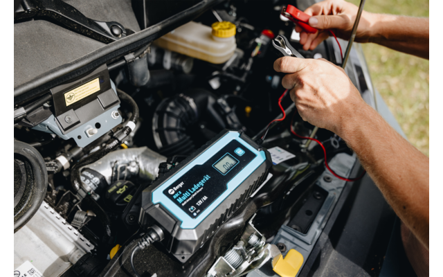 Handen houden rode en zwarte kabelklemmen vast, aangesloten op een zwart-blauw elektronisch apparaat met display in de motorruimte van een auto; motoronderdelen en een gele koelvloeistofdop zichtbaar