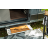 Porte ouverte d'un van aménagé, marche métallique avec paillasson portant le texte "this is us! OUR LIFE OUR ADVENTURE OUR HOME", partie du sol intérieur, herbe à l'extérieur, tapis d'extérieur à motifs et pied de table avec plante en pot