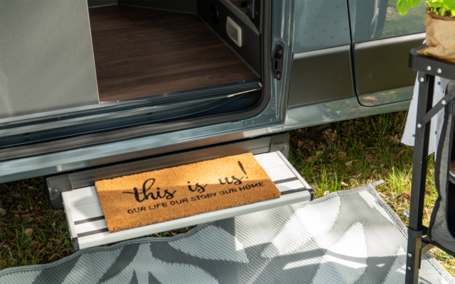 Porte ouverte d'un van aménagé, marche métallique avec paillasson portant le texte "this is us! OUR LIFE OUR ADVENTURE OUR HOME", partie du sol intérieur, herbe à l'extérieur, tapis d'extérieur à motifs et pied de table avec plante en pot