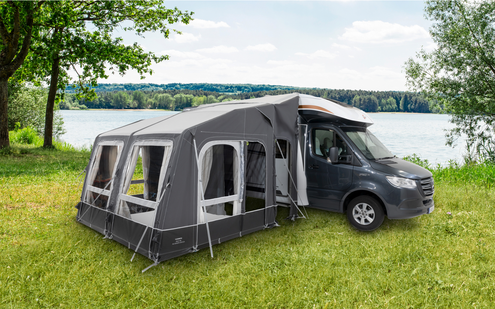 Camping-car noir avec annexe grise sur une prairie verte près d'un lac sous un ciel bleu