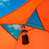 Intérieur d'une tente orange avec des ouvertures triangulaires en résille laissant voir le ciel bleu et des nuages; une lanterne de camping noire pend au centre