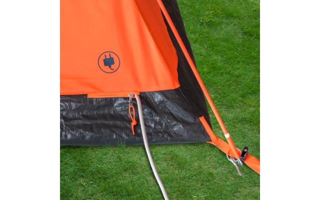 Coin d'une tente orange sur de l'herbe verte, fond de tente noir, arceau orange, câble blanc sortant de la tente, hauban orange avec piquet et mousqueton, pictogramme de prise noir sur la toile