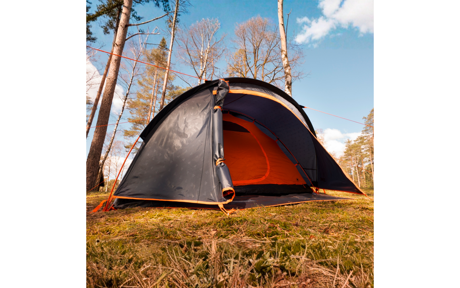 Tente gris foncé, entrée ouverte et intérieur orange, posée sur un sol forestier herbeux entre des arbres fins sous un ciel bleu avec des nuages