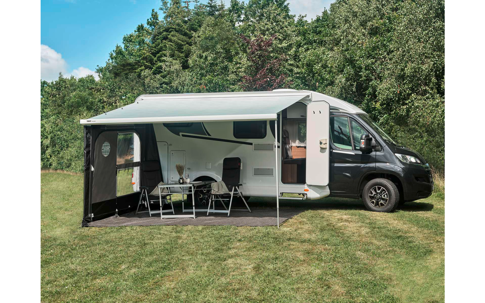 Weißes Wohnmobil mit ausgefahrener Markise, davor Tisch mit Stühlen und kleinem Gegenstand, geöffnete Seitentür, Rasen und Bäume im Hintergrund, blauer Himmel