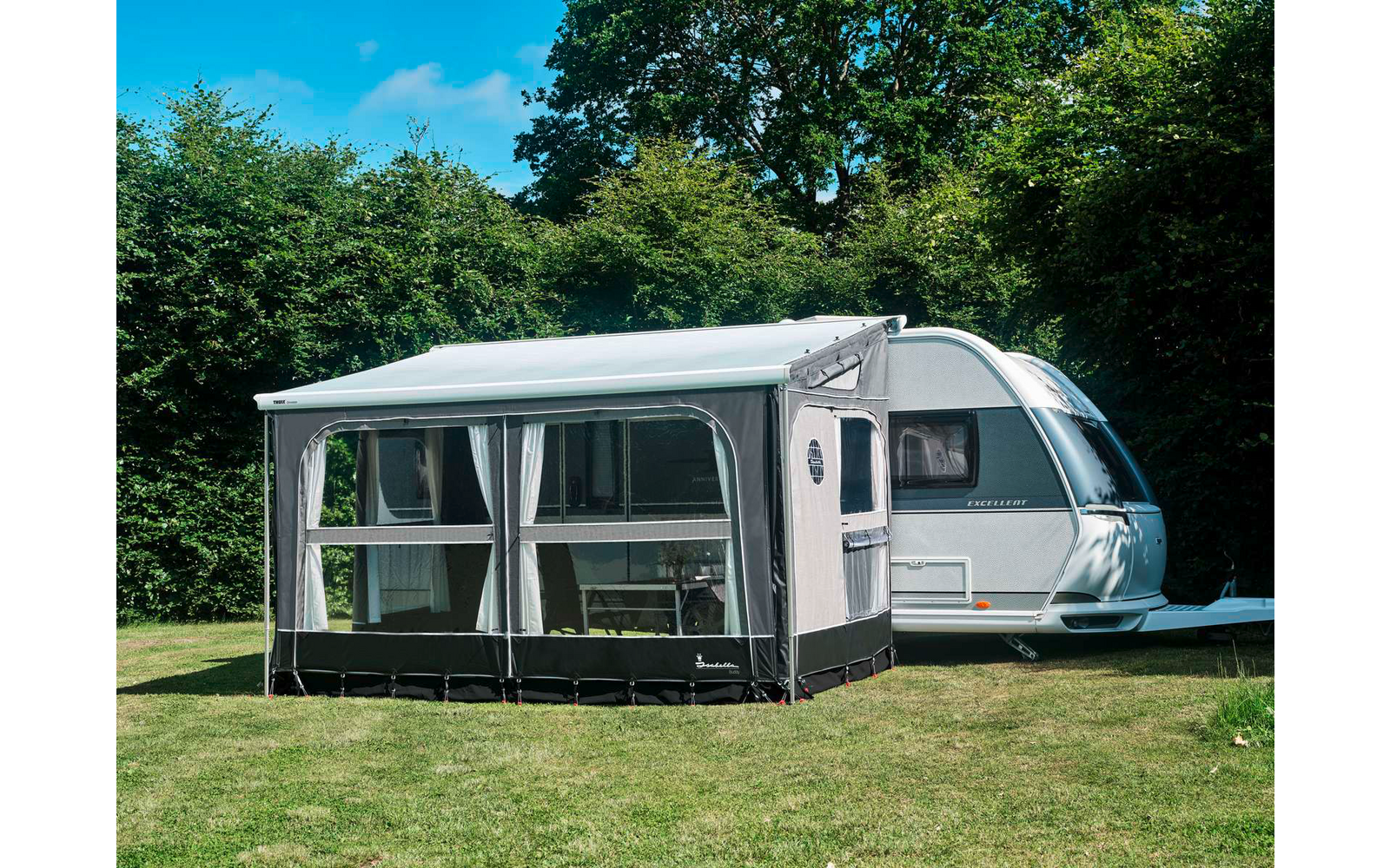 Weißer Wohnwagen mit grauem Vorzelt mit Fenstern und Vorhängen auf einer Wiese, im Hintergrund Bäume und blauer Himmel
