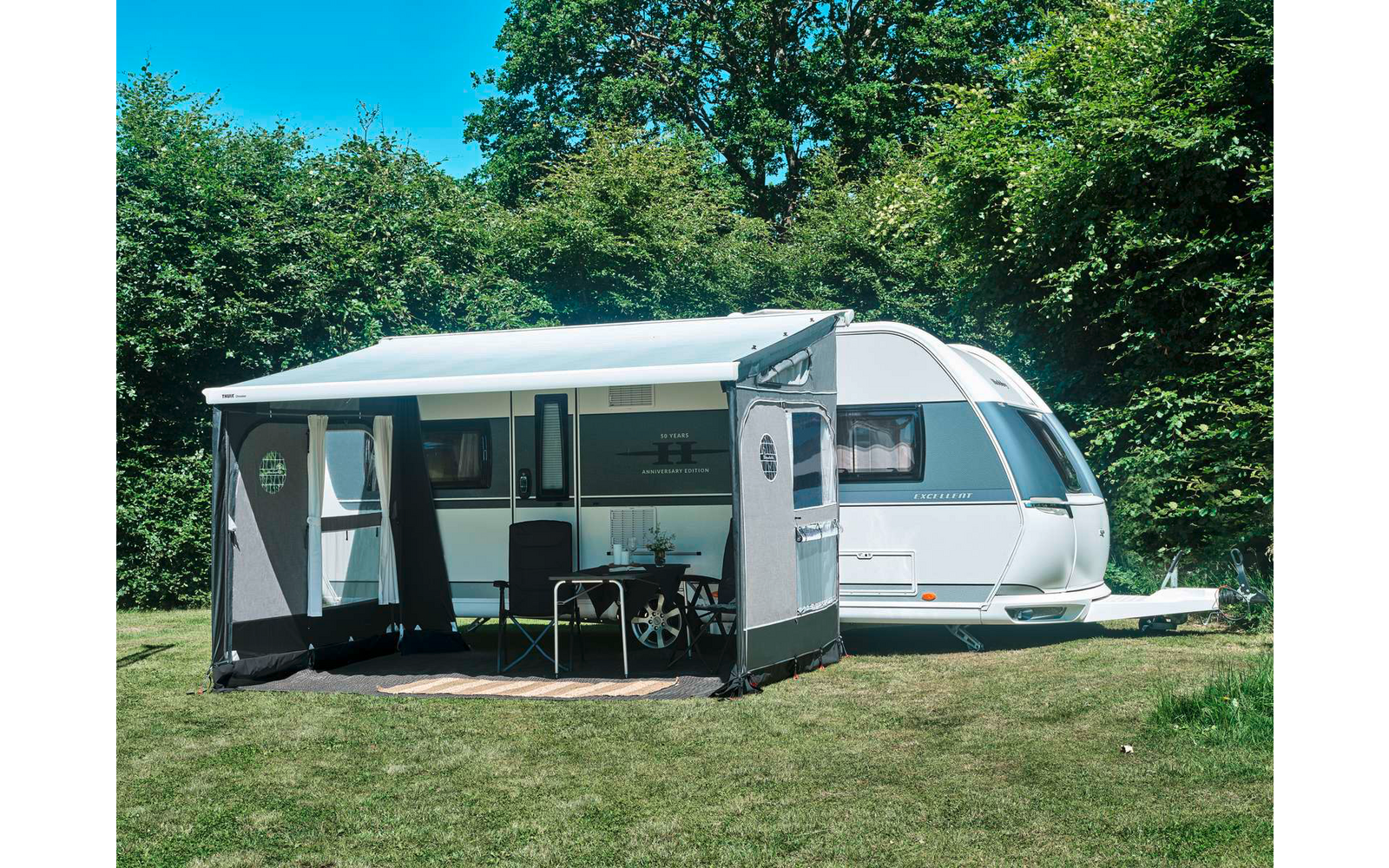 Weißer Wohnwagen auf Rasen mit angebautem grauem Vorzelt; unter dem Vorzelt ein Tisch, zwei Stühle und Gegenstände auf dem Tisch; im Hintergrund Bäume und blauer Himmel