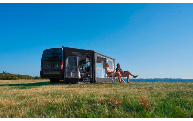 Schwarzer Campingbus mit ausgefahrener Markise, zwei Personen auf Campingstühlen auf einer Wiese, Meer und blauer Himmel im Hintergrund