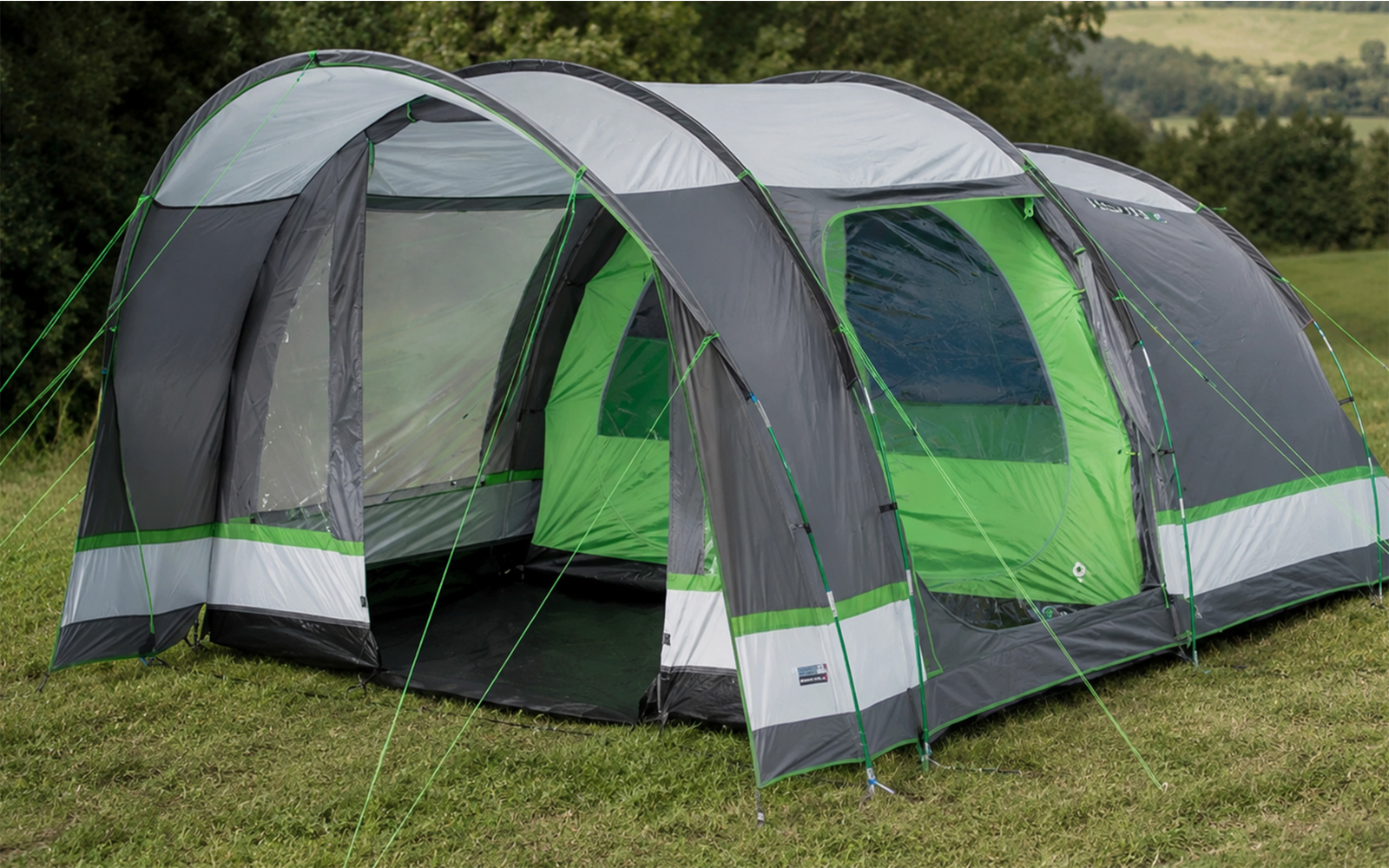 Large gray and green tunnel-style tent with open entrance, side windows, and guy lines staked into grass; treeline in the background