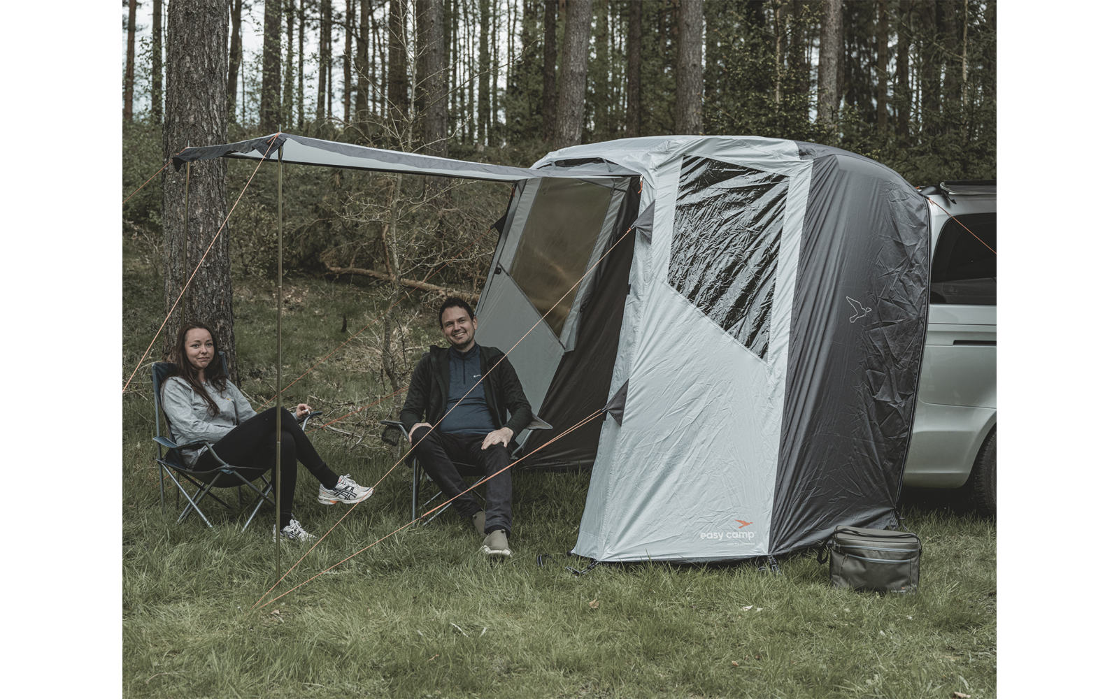 Zwei Personen sitzen auf Campingstühlen neben einem Auto mit angebrachtem Zelt in einem Wald