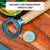 Metallflaschenöffner mit Holzgriff auf Holzuntergrund, daneben ein umgedrehter Kronkorken und eine grüne Verpackung mit Aufkleber 'GEEIGNET FÜR KRONKORKEN-VERSCHLÜSSE'