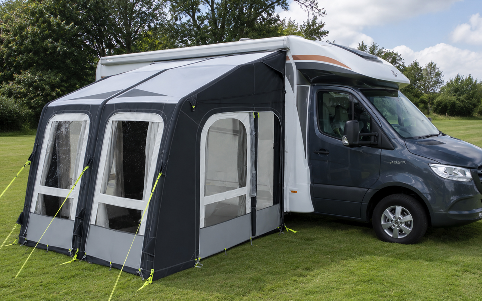 Rechts een grijze camper met een aan de zijkant bevestigd donkergrijze voortent met drie grote ramen, groen gras op de voorgrond, bomen op de achtergrond en een bewolkte lucht