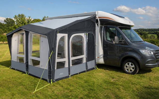 Gray awning with window panels attached to a light gray motorhome on a green lawn, trees in the background and a blue sky with scattered clouds