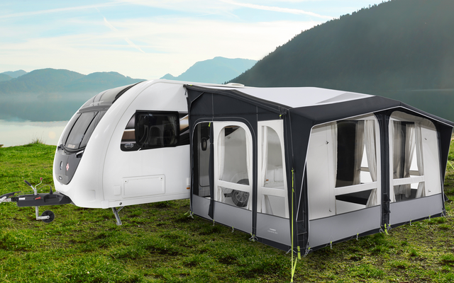 Weißer Wohnwagen mit angebautem, grauem Vorzelt auf einer grünen Wiese am See, Berge im Hintergrund