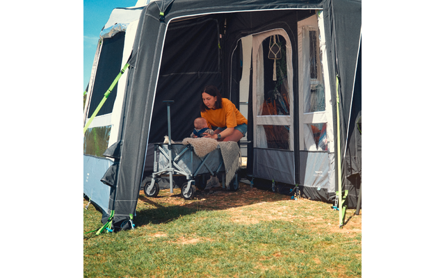 An adult sits in the entrance of a camping awning and leans toward a baby in a stroller