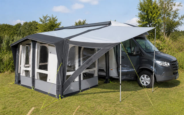 Autocaravana gris con una carpa/tienda gris-blanca fijada al lateral sobre césped; tensores amarillos y postes metálicos; árboles y cielo azul con pocas nubes en el fondo