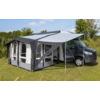 Autocaravana gris con una carpa/tienda gris-blanca fijada al lateral sobre césped; tensores amarillos y postes metálicos; árboles y cielo azul con pocas nubes en el fondo