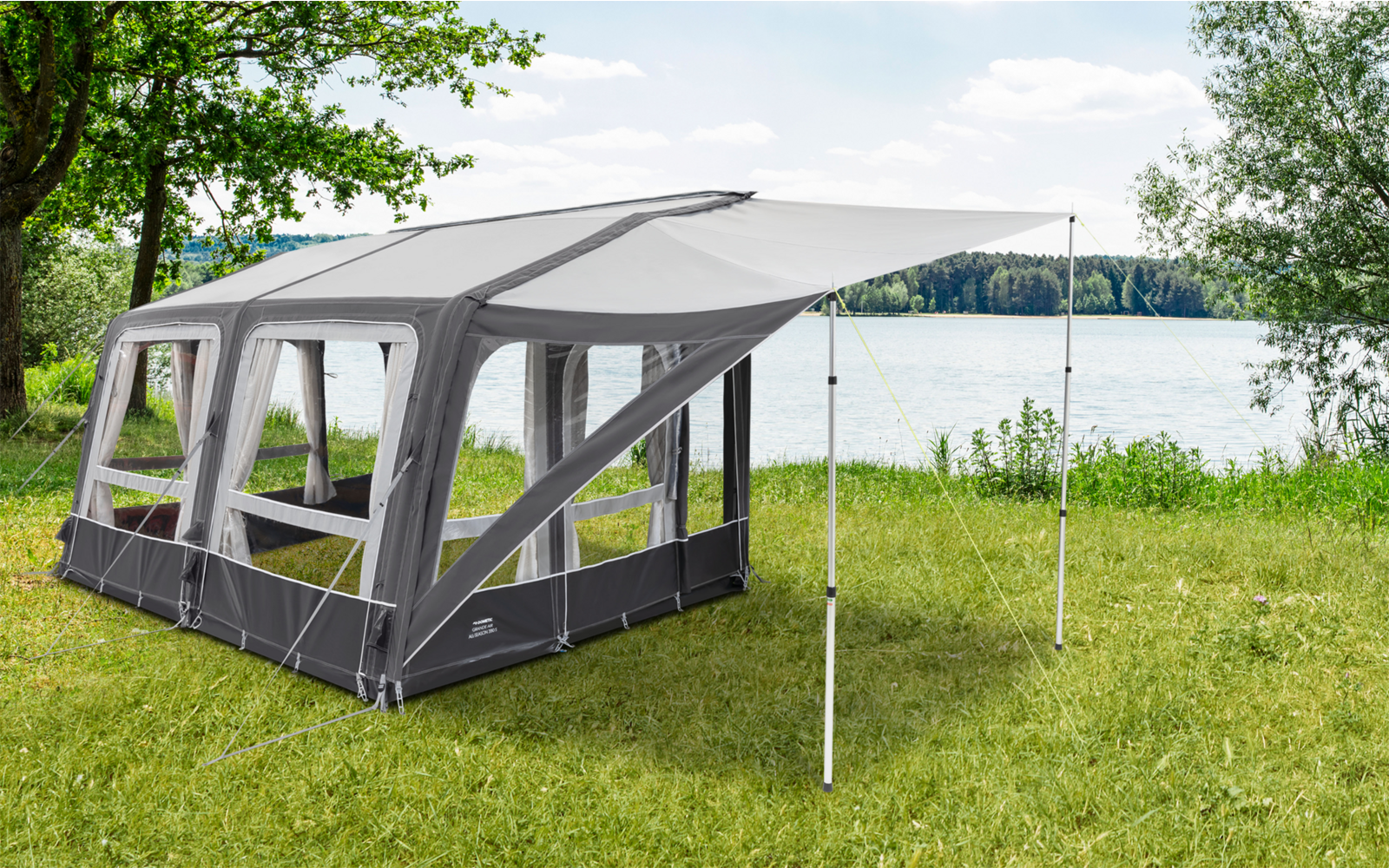 Ein graues und weißes Zelt mit großen Fenstern, aufgestellt auf einer grünen Wiese neben einem See