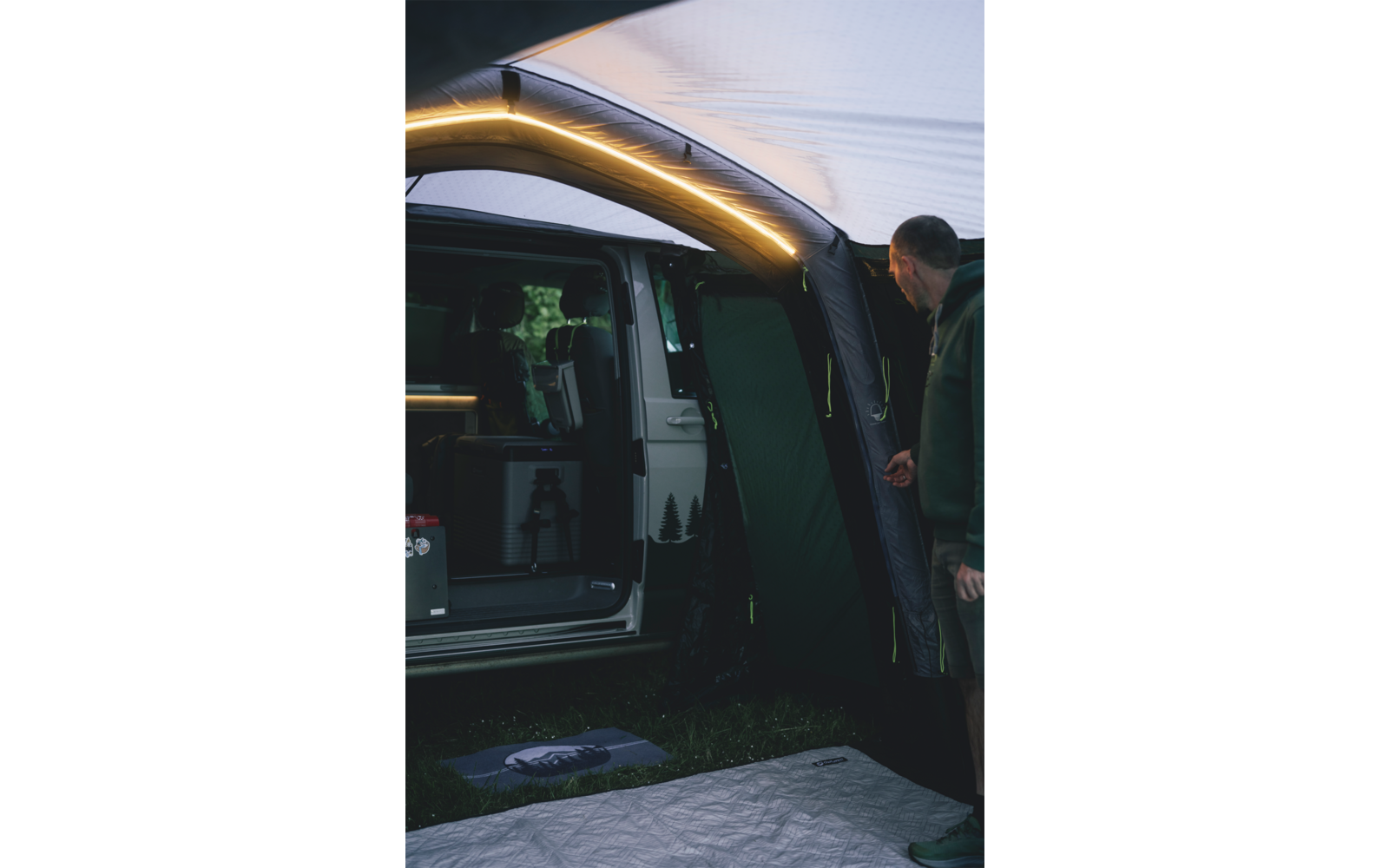 Geöffneter Van mit angebrachtem Vorzelt und beleuchteter Leiste; ein Mann steht rechts am Vorzelt; Sitze und Ausrüstung im Innenraum sichtbar; eine Matte liegt auf dem Gras