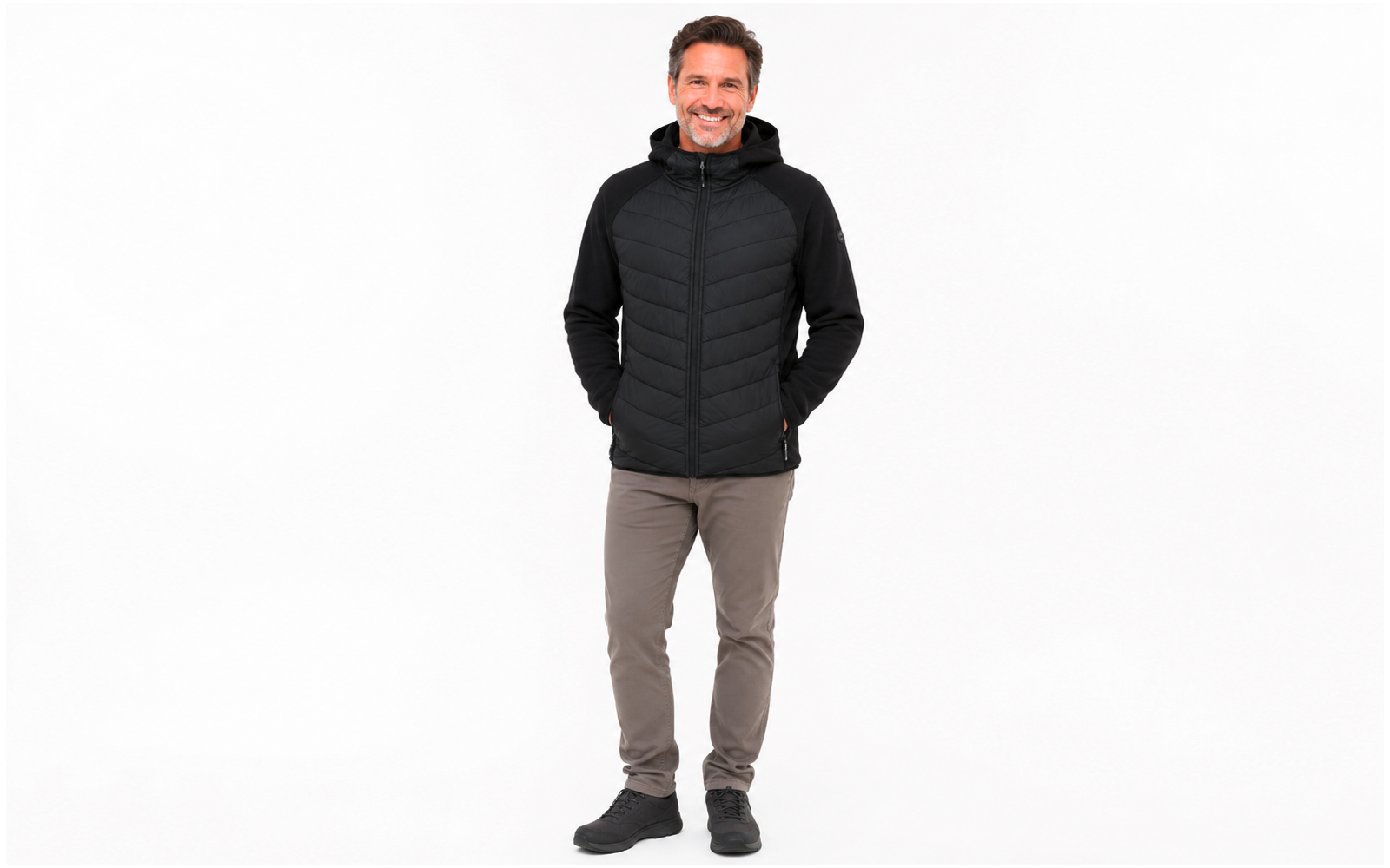 Man in a black quilted jacket with hood, gray pants and black shoes, standing against a white background, hands in pockets, smiling