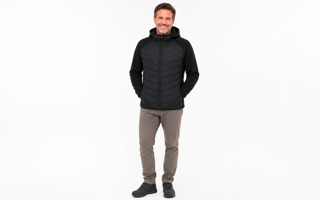 Man in a black quilted jacket with hood, gray pants and black shoes, standing against a white background, hands in pockets, smiling