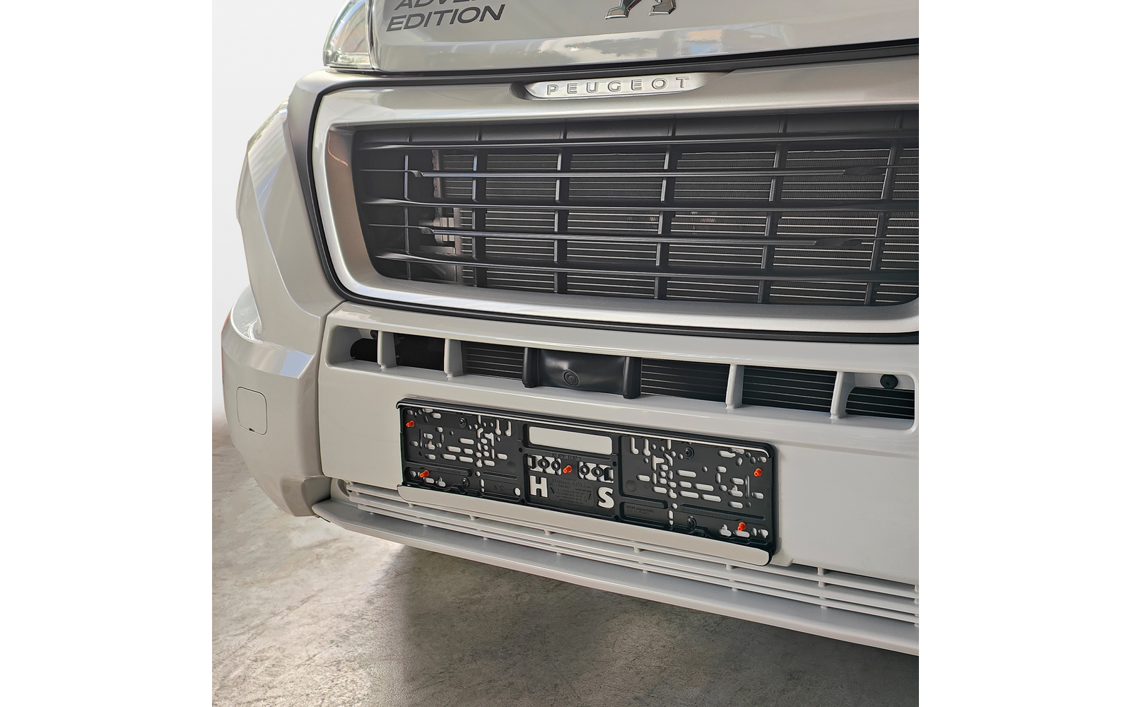 Nahaufnahme der vorderen Fahrzeugpartie eines hellen Peugeot mit schwarzem Kühlergrill, silbernem Peugeot-Schriftzug, leerem Kennzeichenhalter und mehreren runden Öffnungen an der Stoßstange