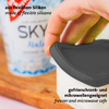 Main pose un couvercle en silicone noir sur un récipient blanc ; emballage flou en arrière-plan ; textes visibles « aus flexiblem Silikon » et « made of flexible silicone » et « gefrierschrank- und mikrowellengeeignet » et « freezer and microwave safe »