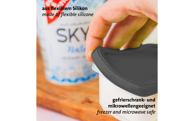 Main pose un couvercle en silicone noir sur un récipient blanc ; emballage flou en arrière-plan ; textes visibles « aus flexiblem Silikon » et « made of flexible silicone » et « gefrierschrank- und mikrowellengeeignet » et « freezer and microwave safe »