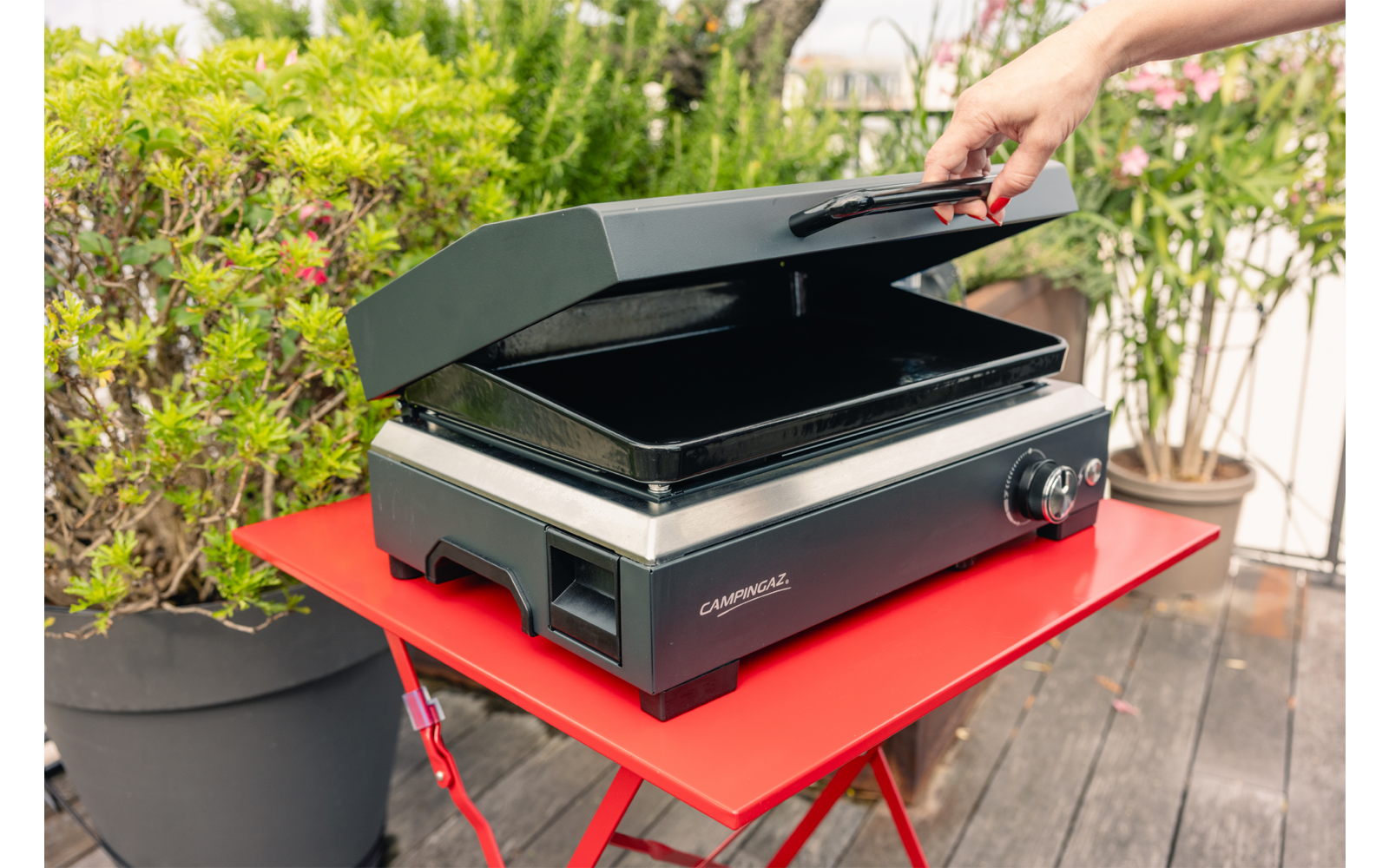 Hand hebt den geöffneten Deckel eines schwarzen Elektrogrills auf einem roten Klapptisch, Topfpflanzen im Hintergrund auf einer Holzterrasse