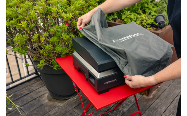 Hände mit roten Fingernägeln ziehen eine graue Stoffhülle von einer schwarzen Schachtel auf einem roten Klapptisch; Pflanze und Holzterrasse im Hintergrund