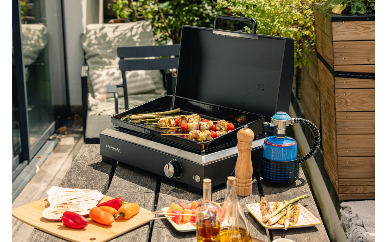 Schwarzer tragbarer Gasgrill mit offenem Deckel und Gemüsespießen auf dem Rost; daneben blauer Gasbehälter, Holz-Pfeffermühle, Schneidebrett mit Fladenbrot und Paprika sowie Ölfläschchen auf einer Balkonterrasse mit Pflanzen und Stuhl
