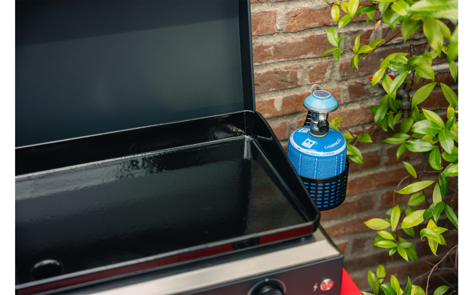 Geöffneter Grill mit leerer schwarzer Grillwanne, angeschlossener blauer Gaskartusche, Backsteinmauer im Hintergrund und grünen Pflanzen rechts