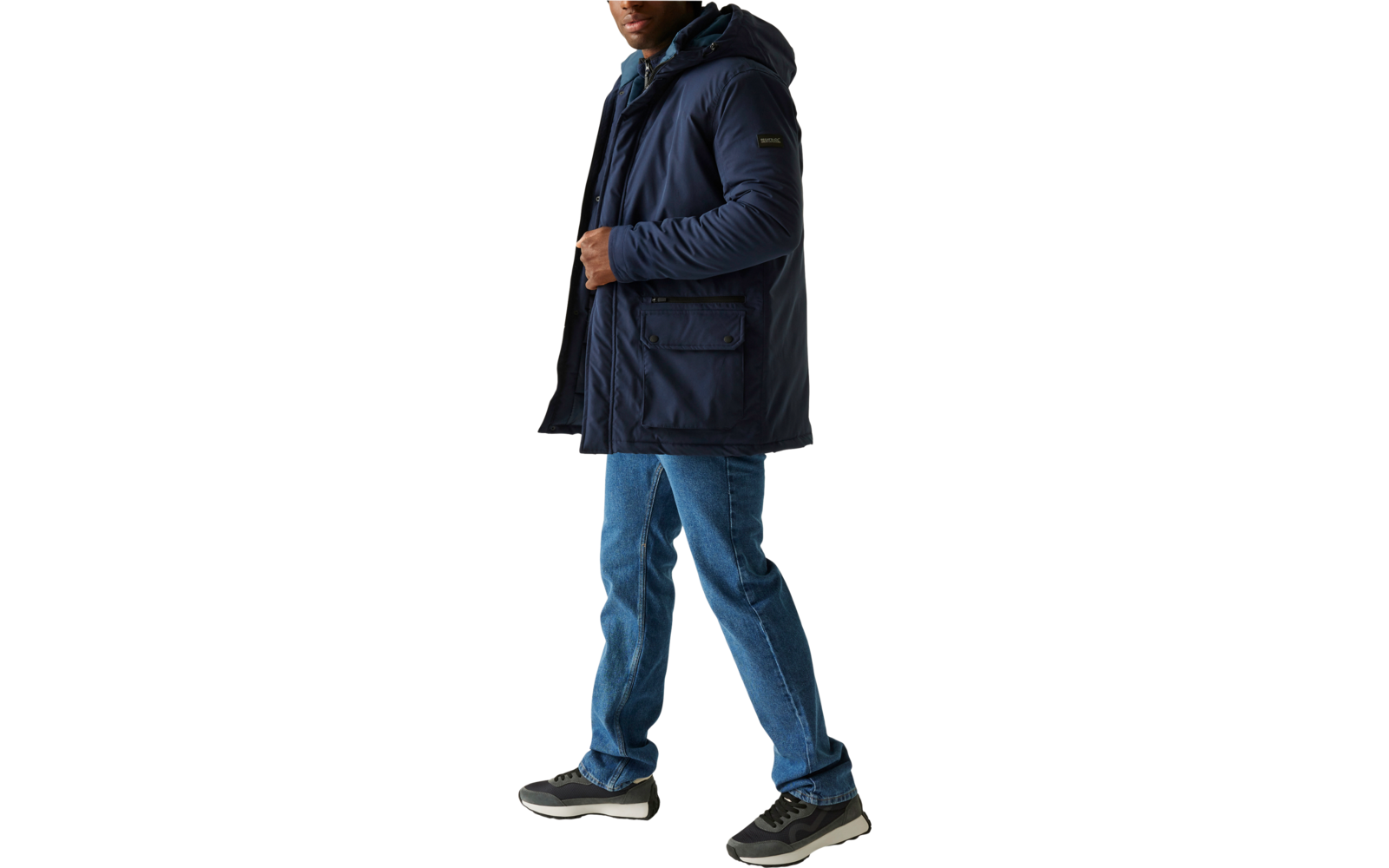 Ein Mann trägt eine blaue Winterjacke, blaue Jeans und schwarze Schuhe, er blickt zur Seite und steht vor einem hellen Hintergrund