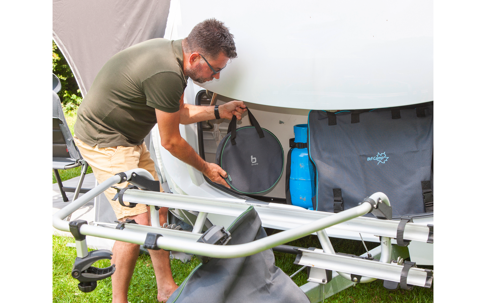 Homme avec lunettes de soleil et T‑shirt vert se penche vers un coffre de rangement ouvert d’une caravane et y place un sac rond noir ; une bouteille de gaz bleue et des sacs gris sont à côté